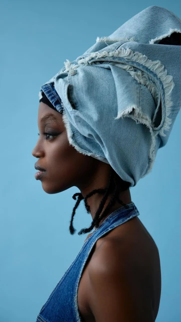 Profile shot of a young Black woman wearing a denim head wrap and halter top against a solid light blue backdrop.