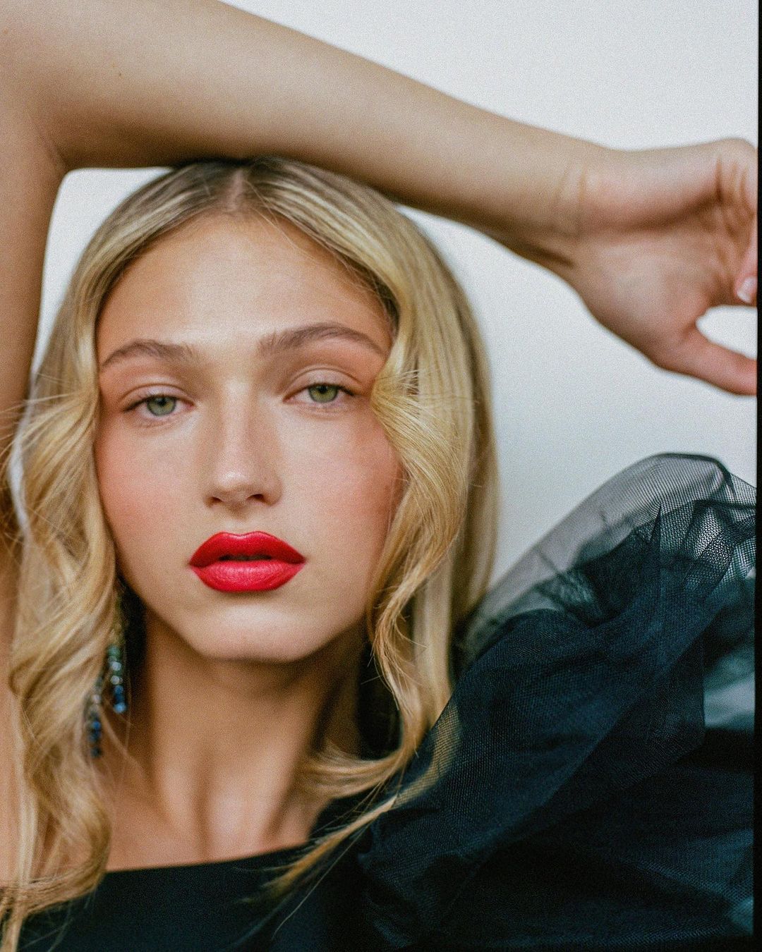 Close-up portrait of a blonde woman with vibrant red lipstick and a dark tulle shoulder garment, photographed by Alvaro Goveia.