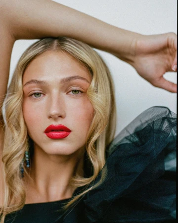Close-up portrait of a blonde woman with vibrant red lipstick and a dark tulle shoulder garment, photographed by Alvaro Goveia.