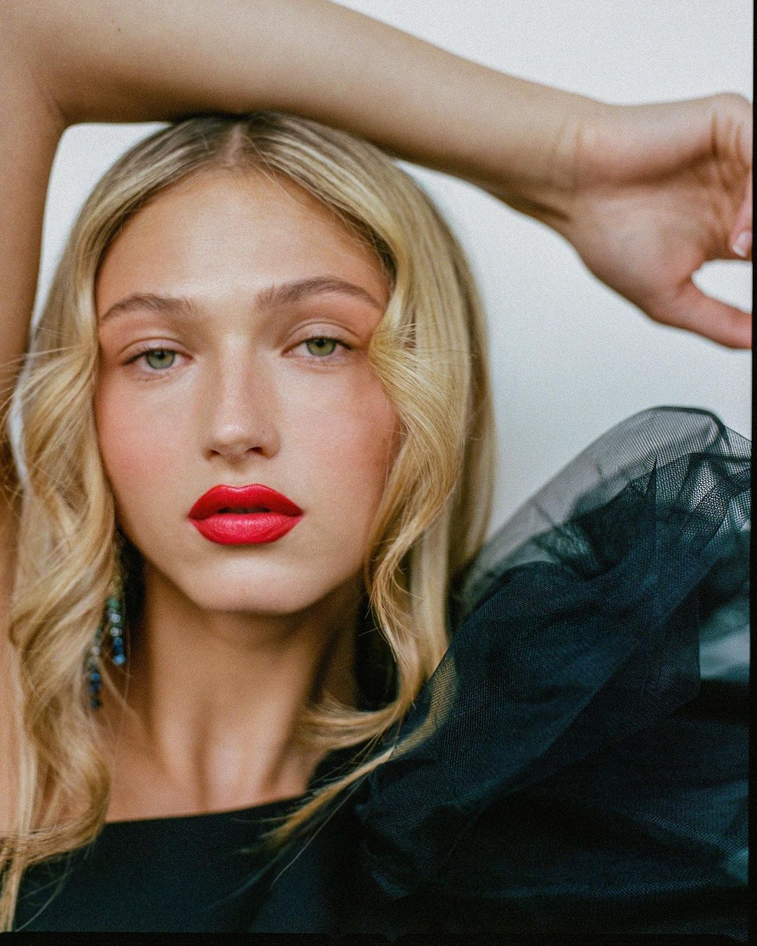 Portrait of a young woman with blonde hair and red lipstick, wearing a black dress, posing in front of a neutral background.