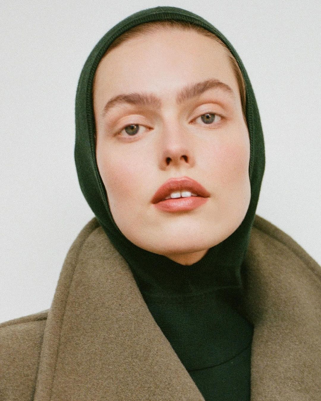 Close-up of a fair-skinned adult woman wearing a dark green balaclava and olive-green coat against a white background in a studio setting.