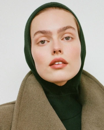 Close-up portrait of a woman wearing a dark green hood and an olive wool coat against a plain background.