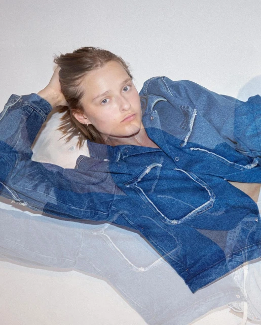 An adult in a denim jacket reclines against a white wall, gazing off-camera with a neutral expression in a portrait-style shot.