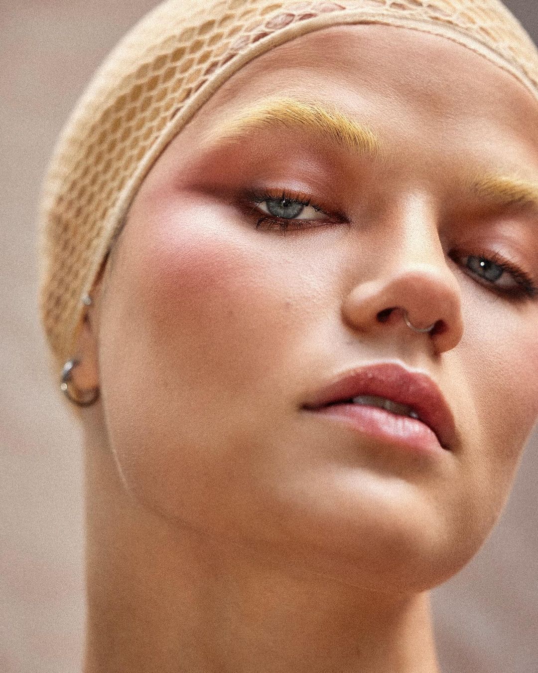 Close-up portrait of a woman with bleached eyebrows and a tan net head covering, featuring soft pink makeup and silver hoop earrings.