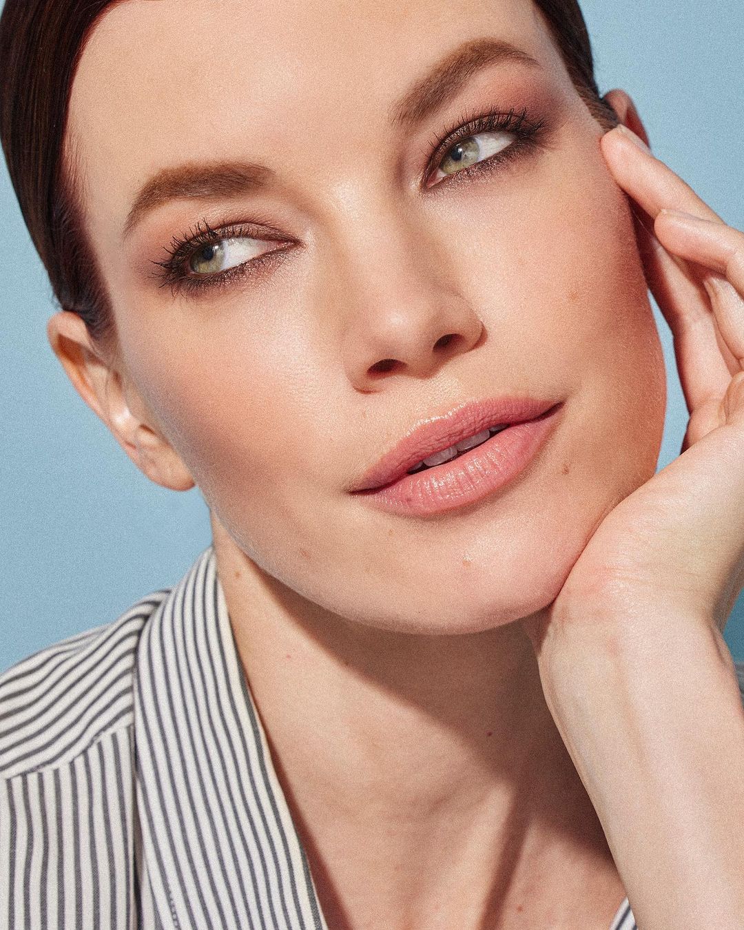 Close-up portrait of an adult woman with fair skin, light eye makeup, and a striped shirt, posing indoors with soft lighting.