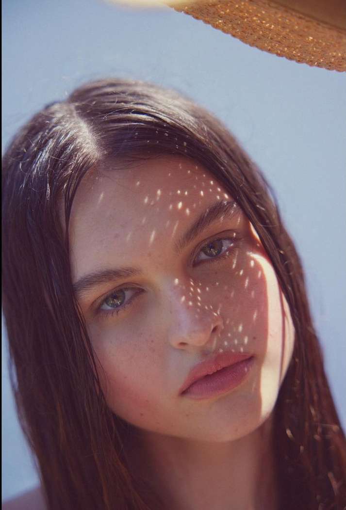 Close-up portrait of a young woman with long brown hair, shadowed face, soft lighting, and a shallow depth of field, against a light blue background.