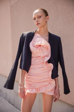 Model in ruffled pink dress and black jacket posing against a textured wall by Alvaro Goveia