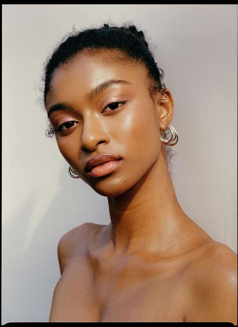 Close-up portrait of a young adult female with dark hair and glowing skin, wearing hoop earrings, set against a neutral background.