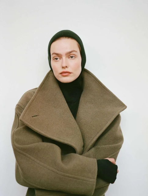 A woman wearing a dark green hat and a wool coat, set against a plain white background. The model gazes directly at the viewer.