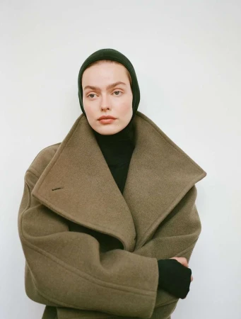 A woman wearing a dark green hat and a wool coat, set against a plain white background. The model gazes directly at the viewer.