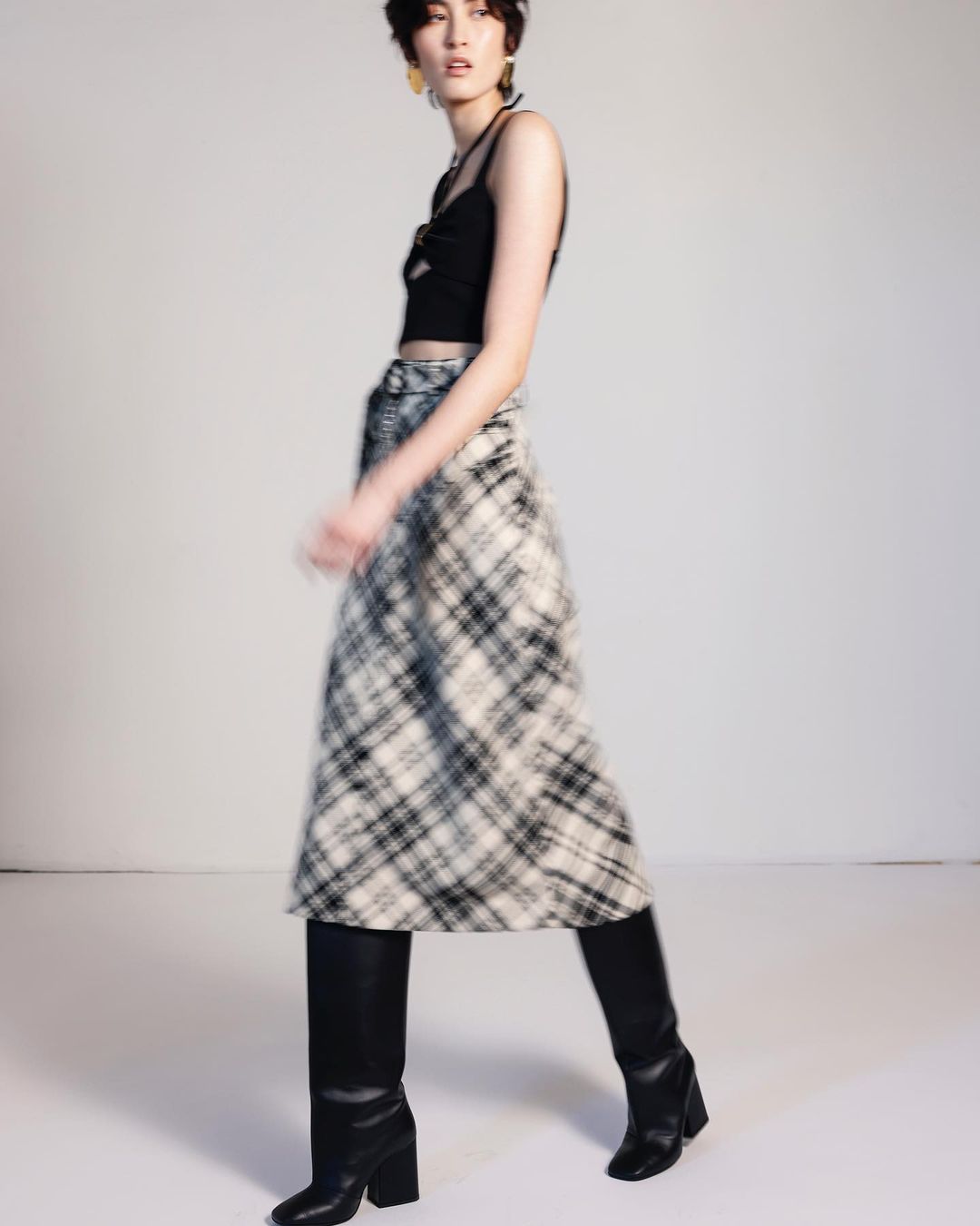 Model wearing a black crop top and plaid midi skirt with black knee-high boots in a studio setting