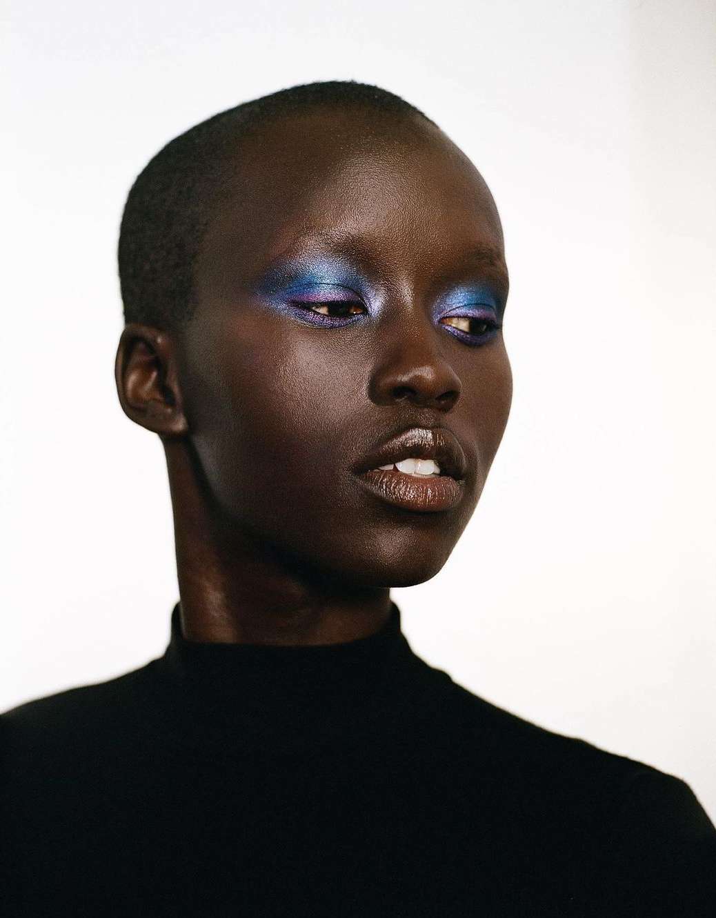 Close-up portrait of a Black woman with blue and purple eye makeup, wearing a black turtleneck sweater against a plain white background.