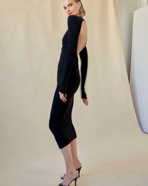 Elegant adult woman with short blonde hair poses in a black, long-sleeved backless midi dress and black high-heeled mules in a studio.