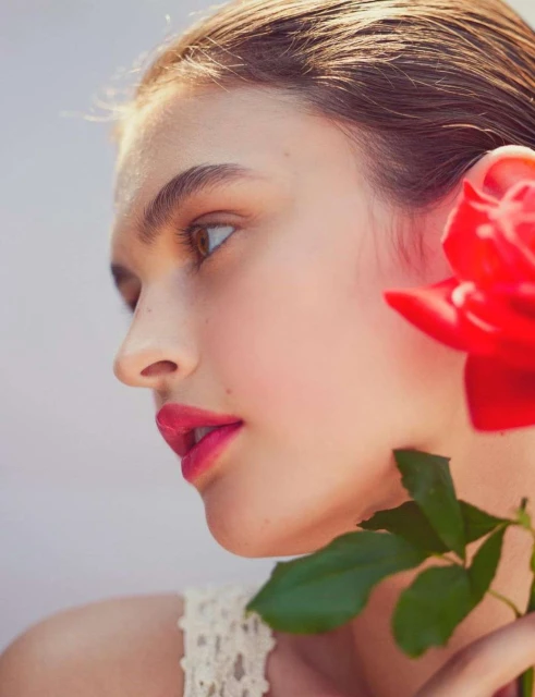 Profile of a woman with fair skin and pink lips, accented by a red rose with green leaves against a soft grey backdrop.