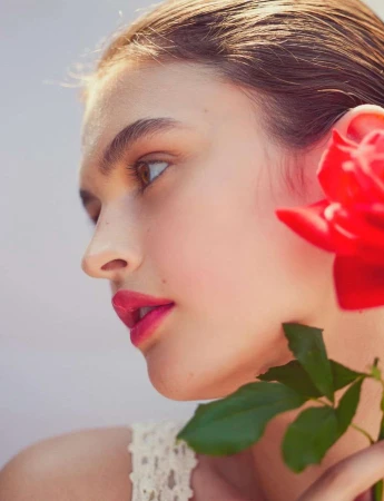 Profile of a woman with fair skin and pink lips, accented by a red rose with green leaves against a soft grey backdrop.