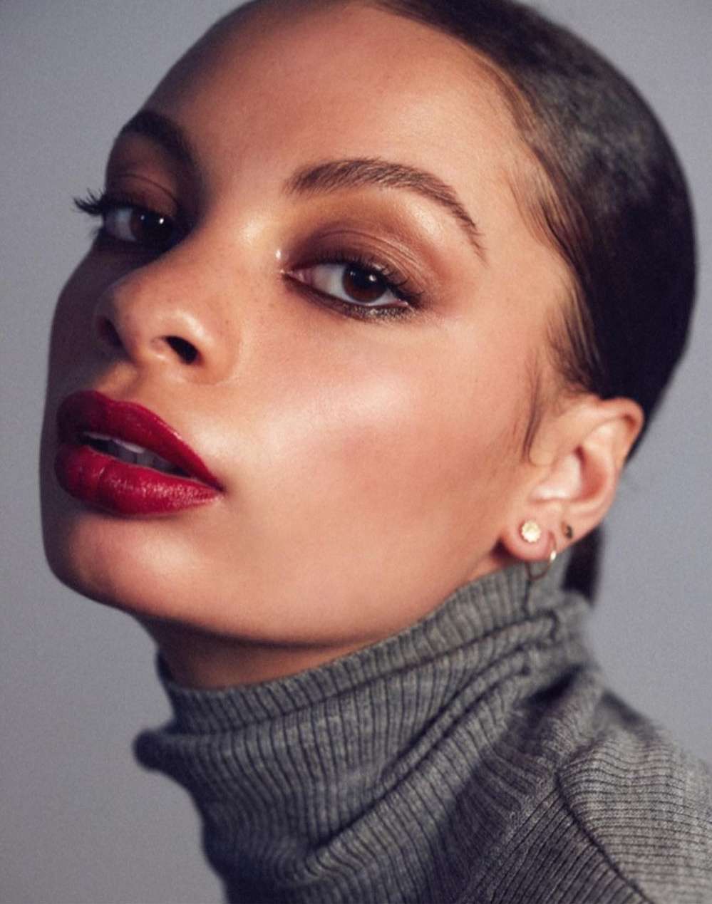 Close-up portrait of an adult woman with slicked-back hair and dark red lipstick, wearing a grey turtleneck sweater against a grey backdrop.