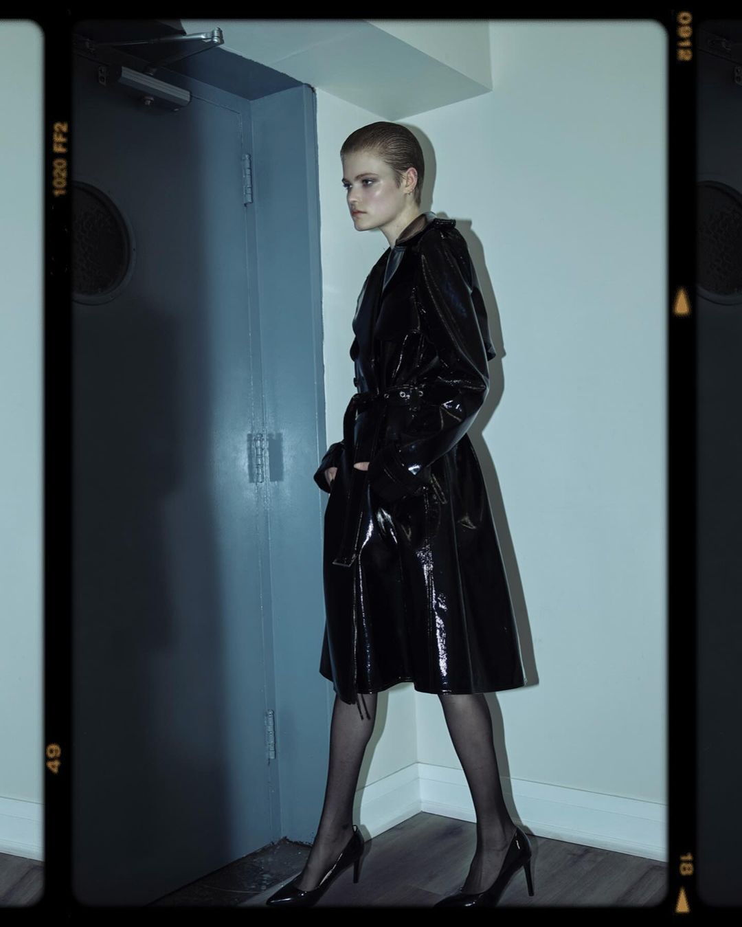 A woman poses in a glossy black trench coat, sheer black stockings, and high heels in an indoor setting with blue walls and doorways.