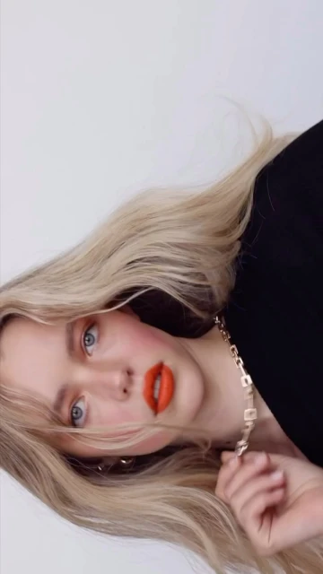 Close-up portrait of a fair-skinned woman with long blonde hair, blue eyes, and orange lipstick wearing a black top and gold chain necklace.