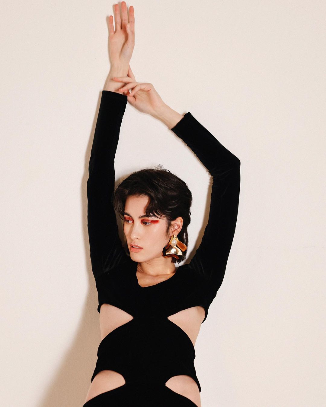 A woman in a black cutout dress poses against a white wall, with her arms raised, showcasing fashion and style in a minimalist setting.