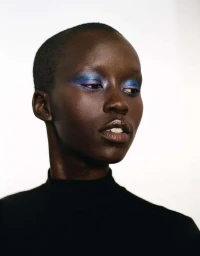 Close-up portrait of a Black woman with a shaved head wearing bold blue eyeshadow and a black turtleneck against a white background.