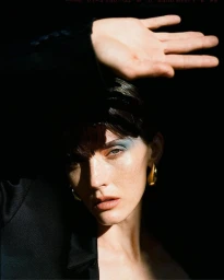 Close-up portrait of a woman with blue eyeshadow and gold earrings holding her hand up against a dark background