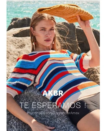 Model in colourful striped knit top holding straw sun hat against rocky beach and ocean background