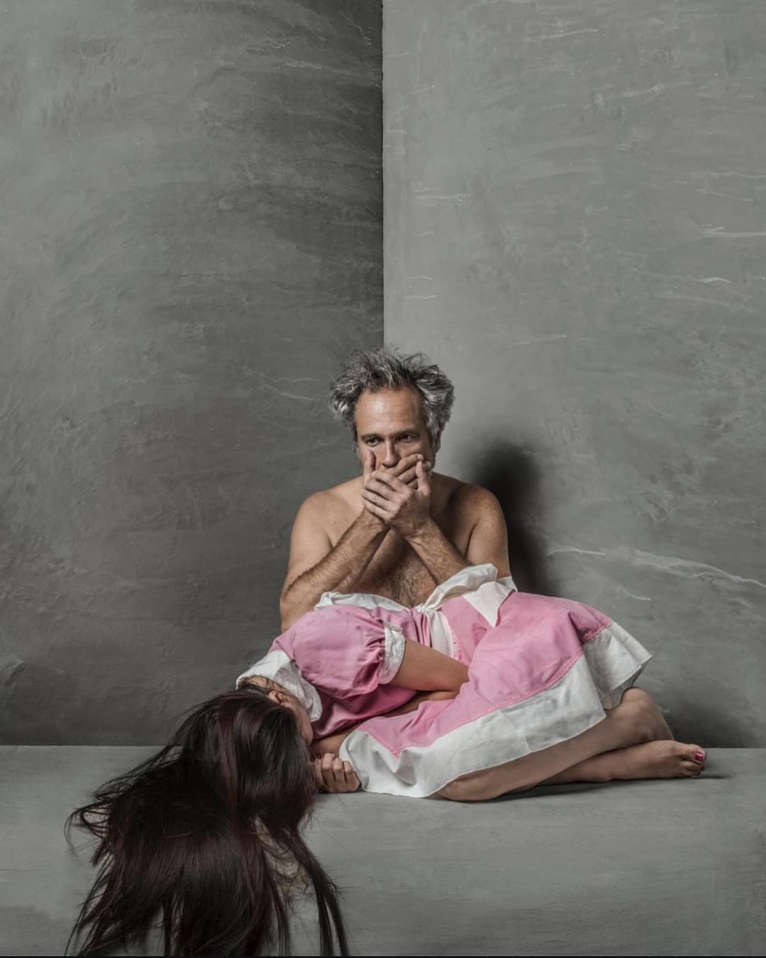 Mature man covering mouth with hands with child lying down in pink dress in indoor studio setting with grey walls.