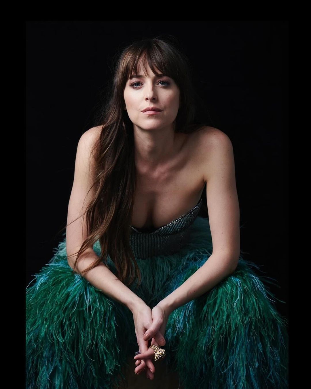 Portrait of a woman with long brown hair, wearing a strapless top and feathered skirt, posing against a dark background in a studio.