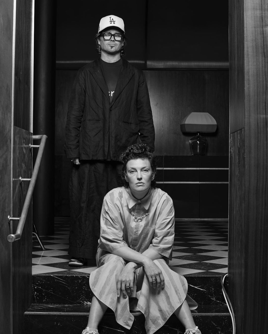 Black-and-white studio shot of a seated woman and standing man wearing casual clothes with serious facial expressions.