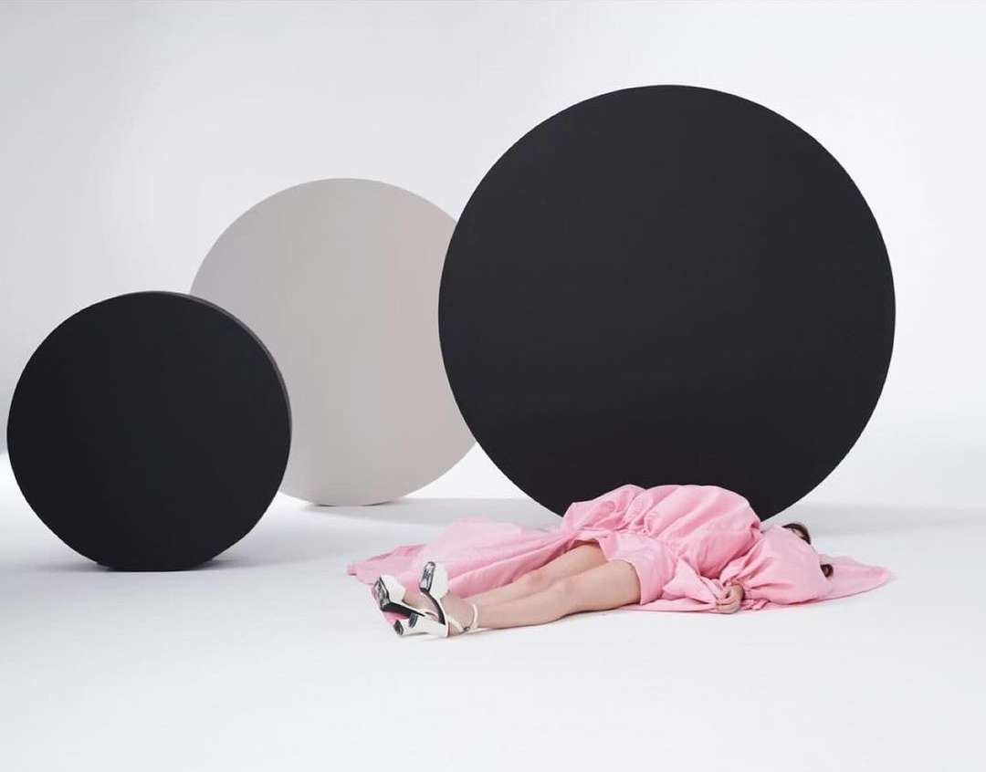 Person in pink dress lying on a white surface with black and white circular backdrops in a fashion studio setting.