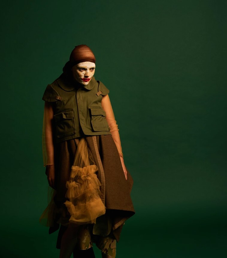 Fashion model poses in olive drab military-style top and brown skirt with tulle accents against a dark green studio backdrop.