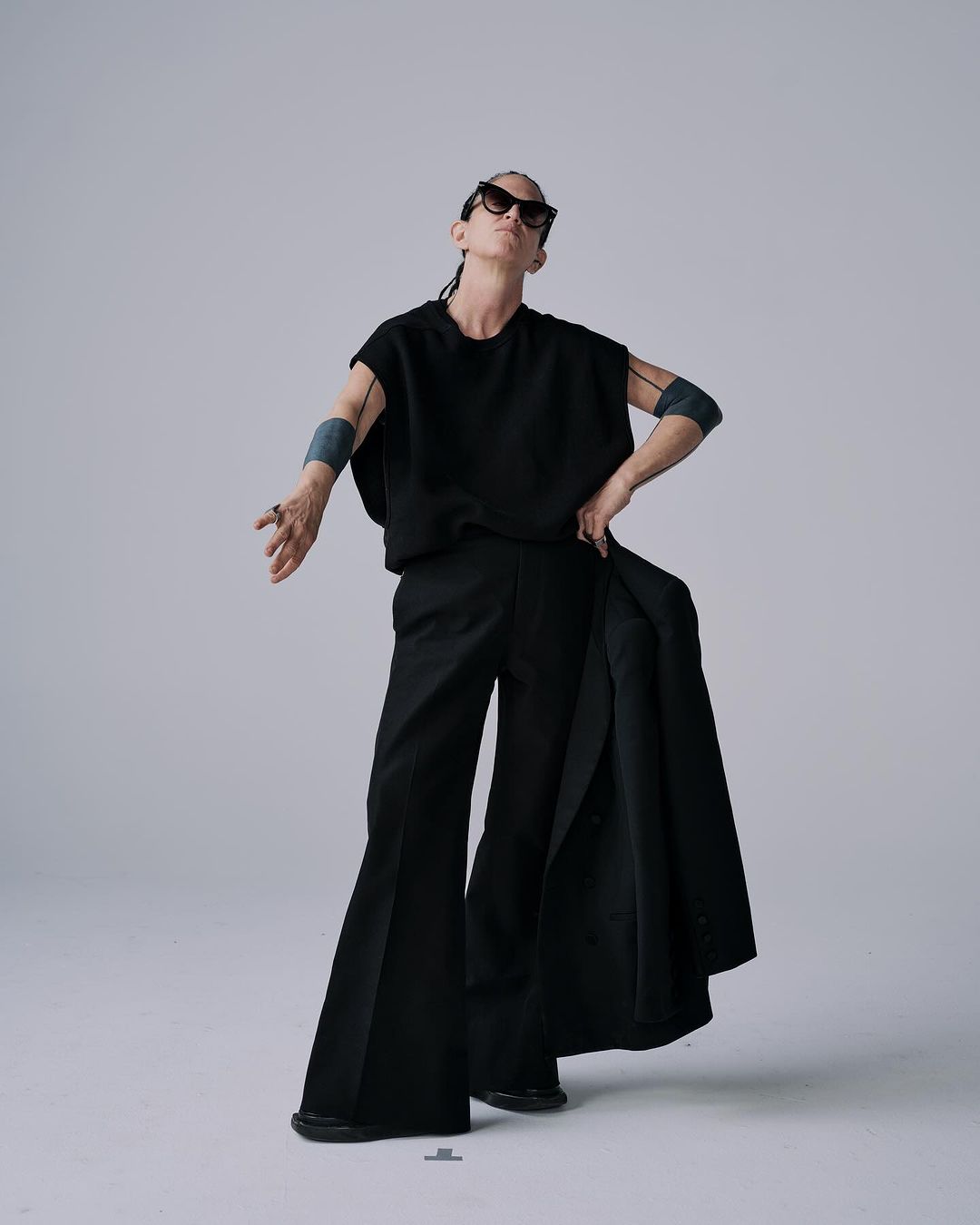 A stylish adult poses in a photo studio, wearing sunglasses, a black top, and wide-leg trousers with a black jacket over the arm.