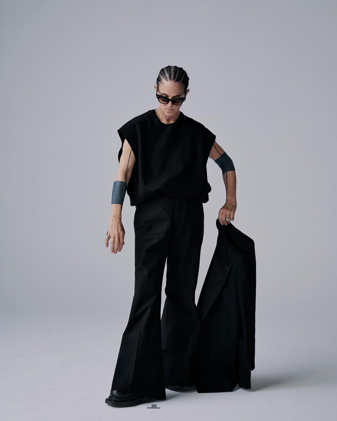 Person in sunglasses posing in black sleeveless top and wide-leg pants, holding a jacket against a grey backdrop in a studio.