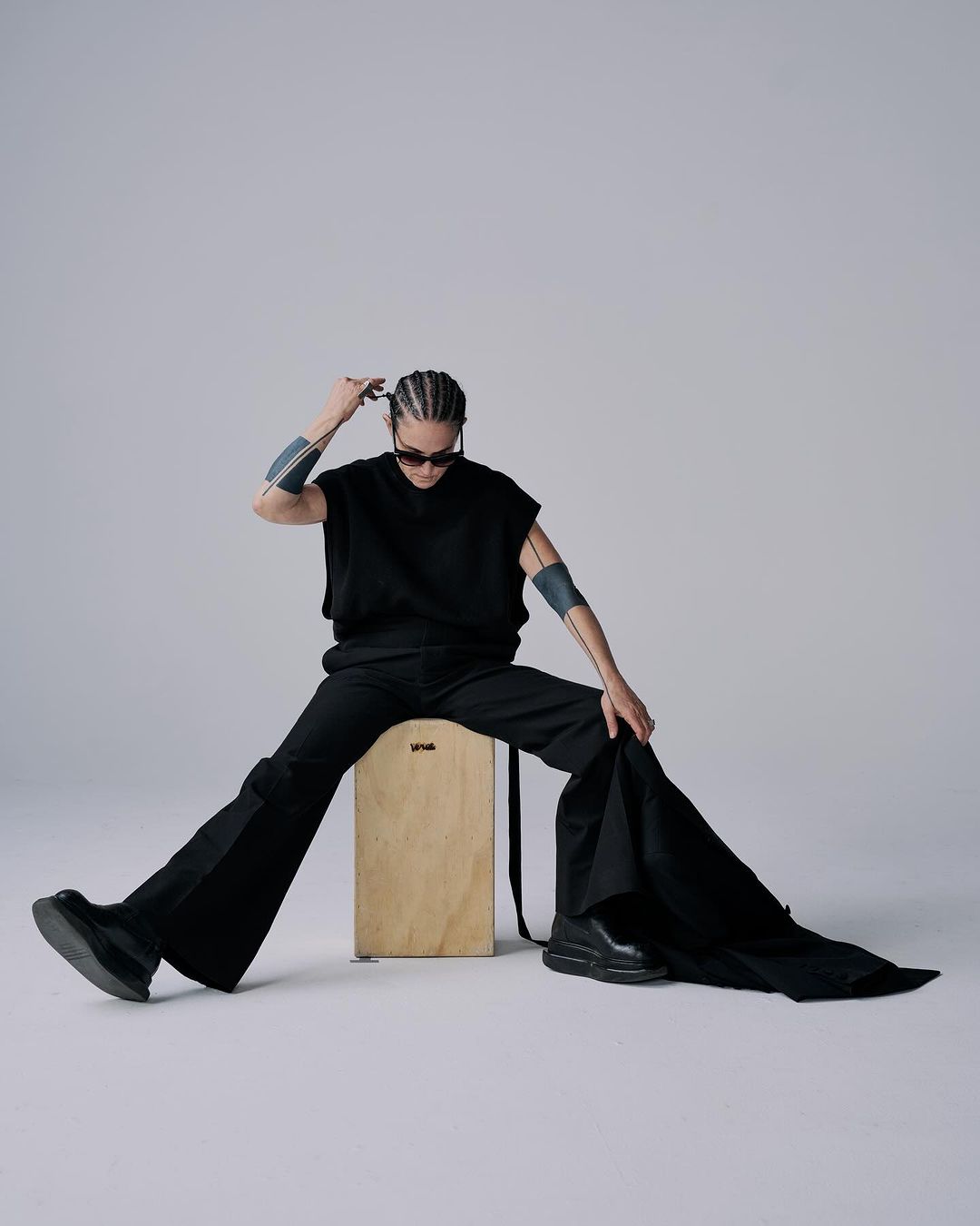 Androgynous person with braided hair sits on a wooden box in black top, pants, sunglasses, and chunky shoes in a light photo studio.