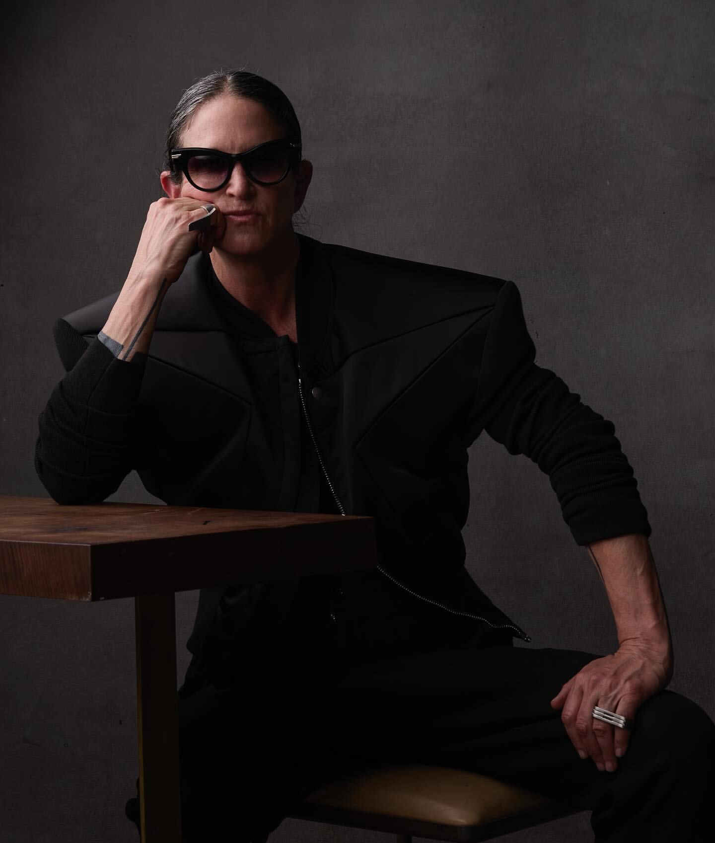 An adult sits at a table wearing sunglasses and a structured black jacket with padded shoulders, shot in a photo studio.
