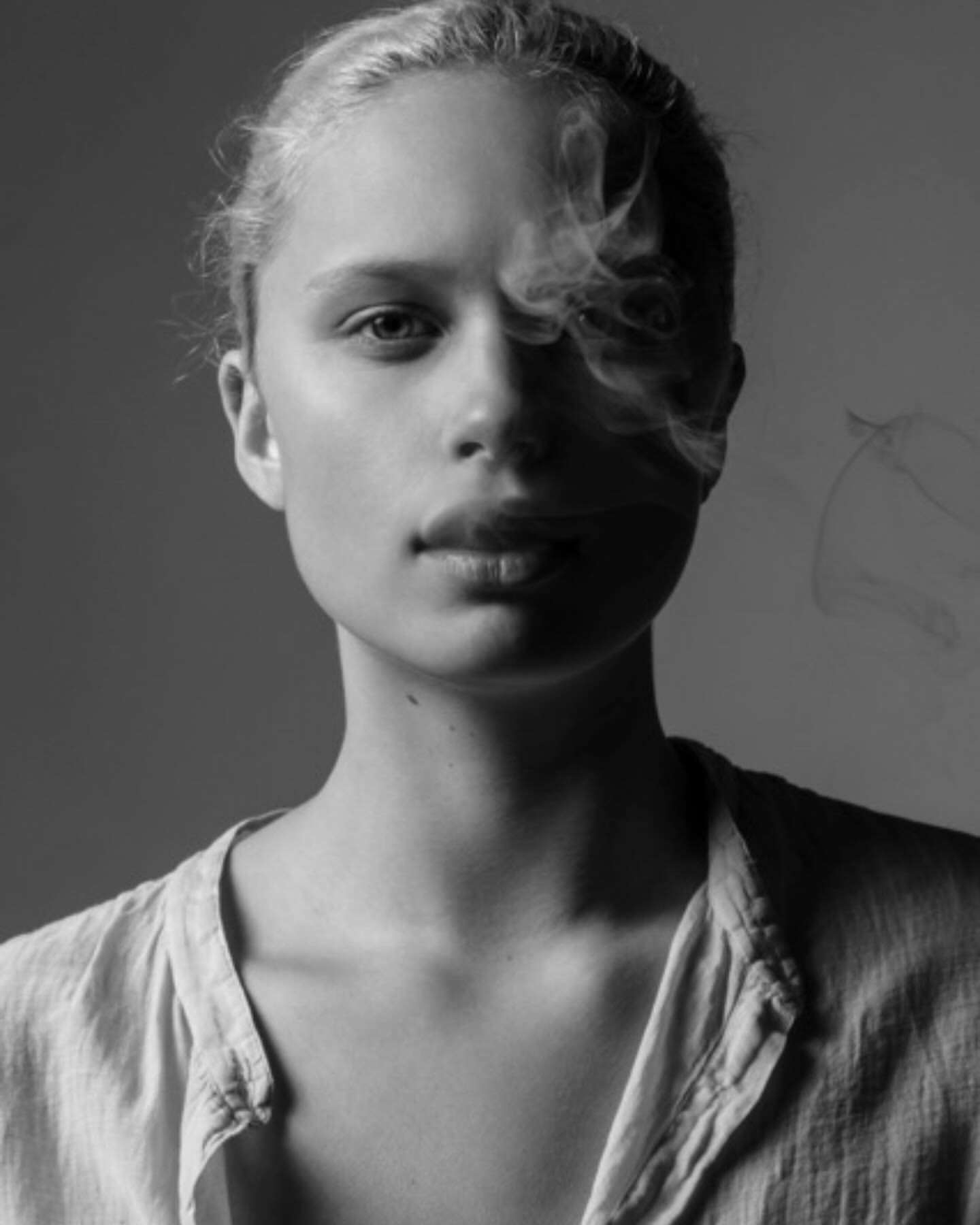 Black and white portrait of a fair-skinned young woman with blonde hair, smoke curling near her face, wearing a linen shirt.