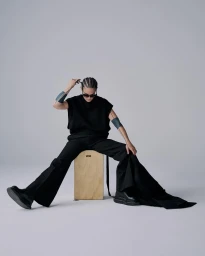 Person in black clothing sitting on a wooden box wearing sunglasses against a white background
