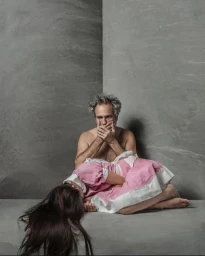 Shirtless man sitting in a corner with a woman in a pink dress curled up in his lap, photographed by Amanda Demme.