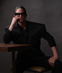 Portrait of a person wearing large sunglasses and a structured black jacket seated at a table against a grey textured studio backdrop.