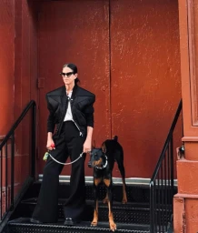 Woman in dark sunglasses, black structured jacket, and wide trousers standing on stairs with a Doberman dog by Amanda Demme
