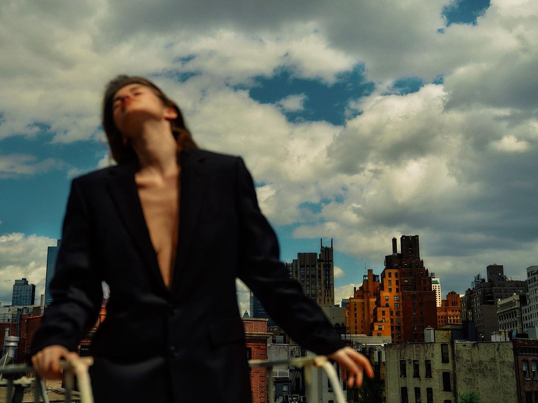 Woman wearing an oversized black blazer outdoors against a city skyline under a dramatic cloudy blue sky.