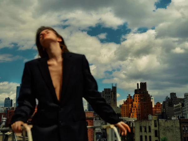 Woman wearing an oversized black blazer outdoors against a city skyline under a dramatic cloudy blue sky.