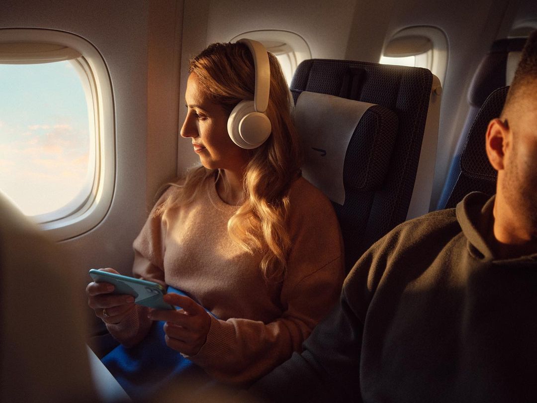 Woman with headphones sits on an airplane using a smartphone, enjoying the view from the window during a sunny, warm flight.