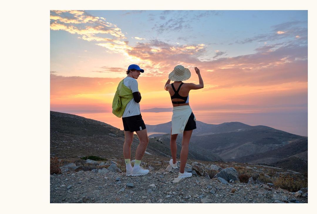 A couple stands atop a scenic mountain, gazing at the sunset. One wears a hat, the other a baseball cap, both in athletic attire.
