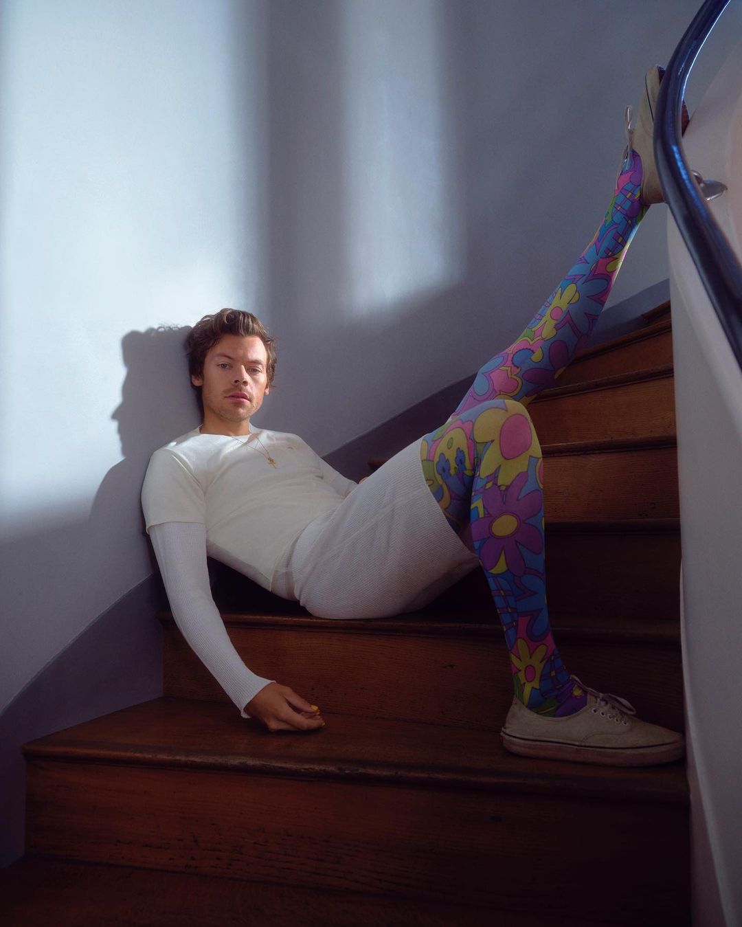 Adult male with floral tights poses sitting on indoor wooden staircase with off-white trainers and a long-sleeve shirt in a studio setting.