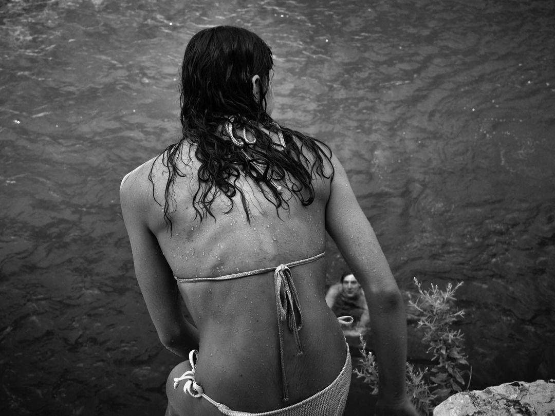 Woman in a bikini stands at the water's edge, her back to the camera, wet hair cascading down her back, monochrome image.