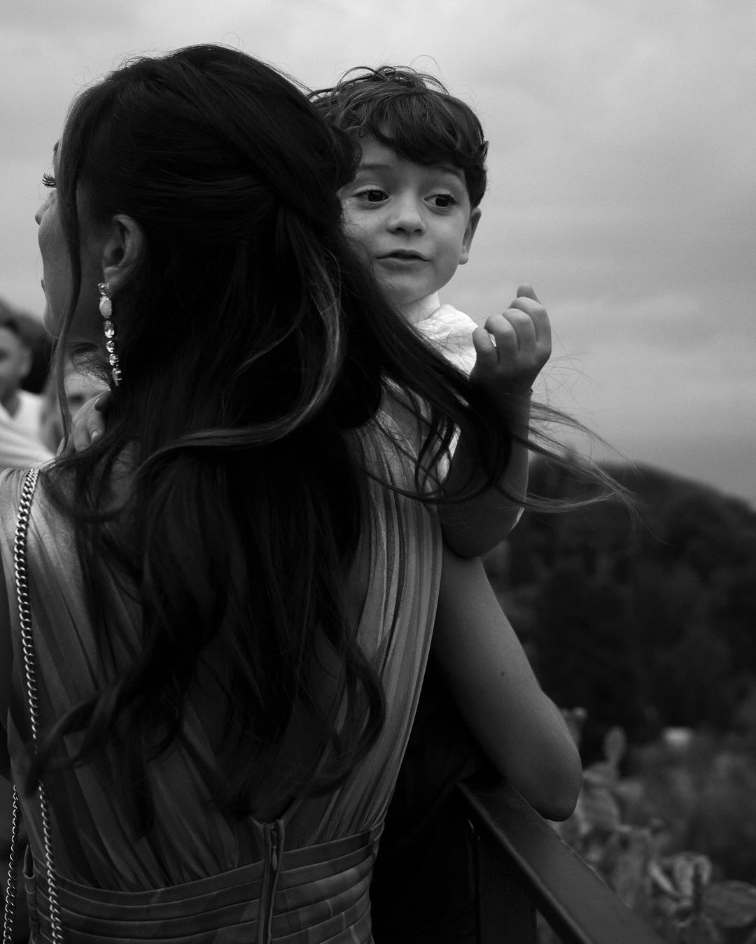 A woman in formal attire holds a child, both gazing into the distance against a soft sky in a black and white outdoor shot.