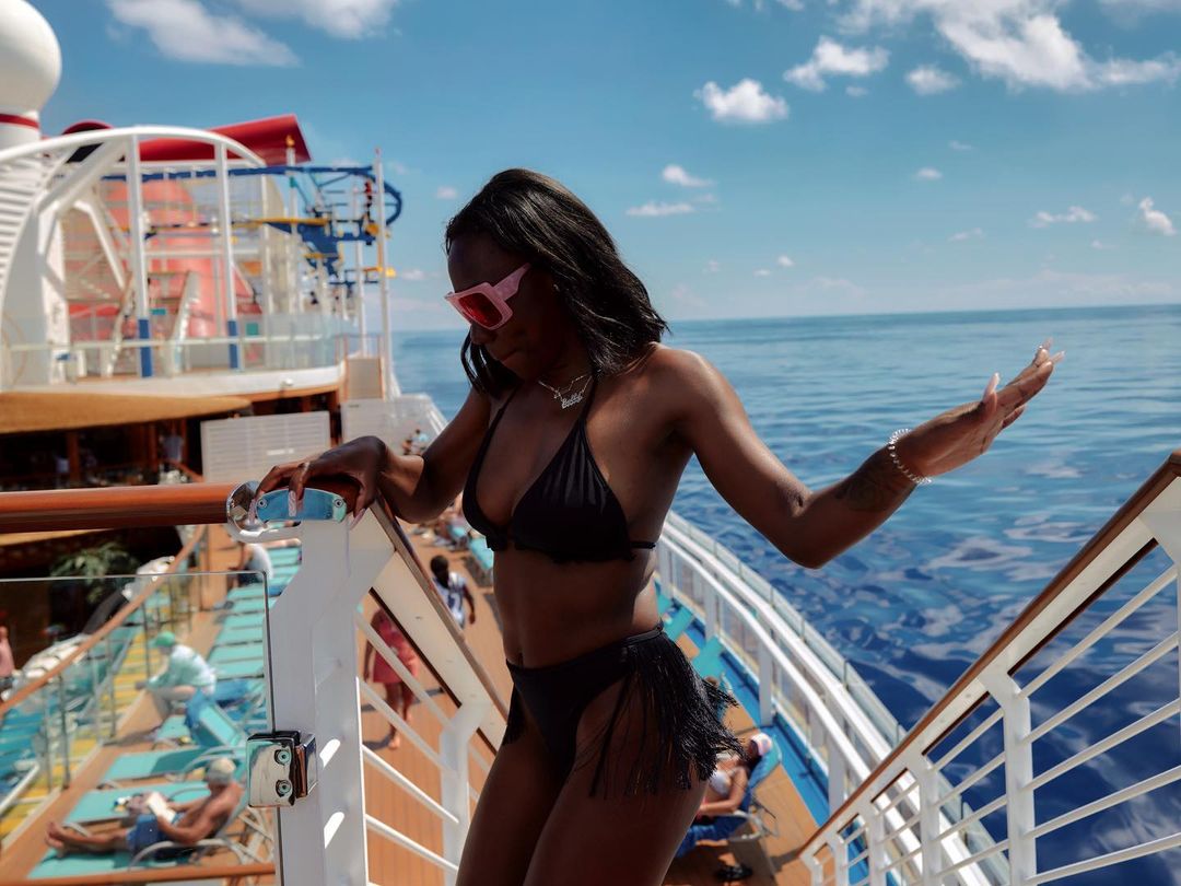 Woman in bikini poses on cruise ship deck with ocean view. She's wearing pink sunglasses on a sunny day while on vacation.