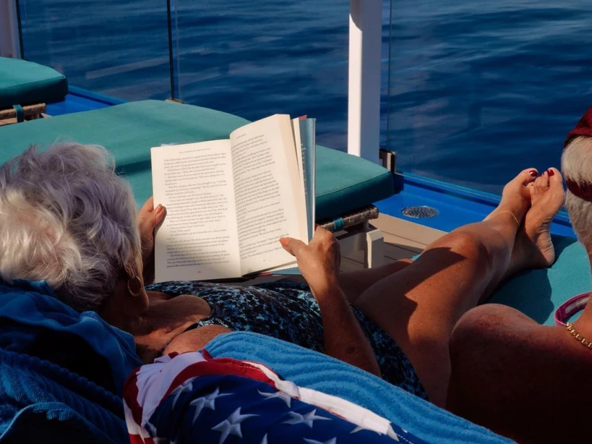 Elderly woman reading a book while sitting on a lounger outdoors in bright sunlight, with the ocean visible in the background.