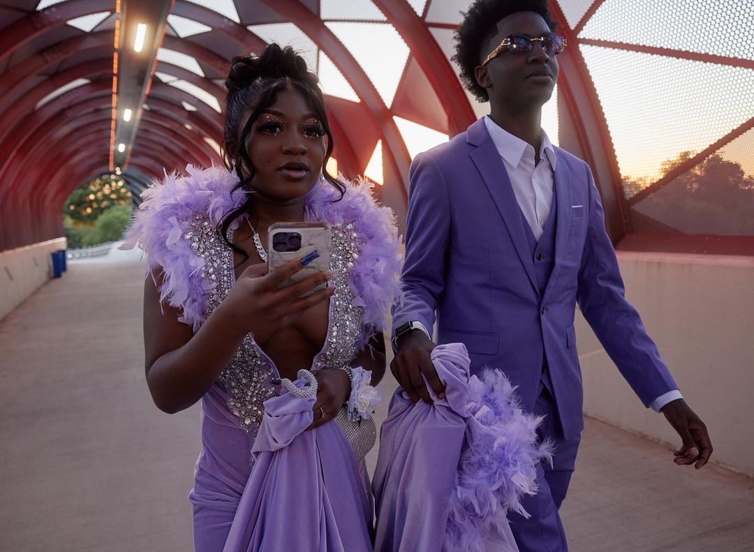A young African-American couple in formal wear walk outdoors on a covered walkway. The young woman holds a smartphone.
